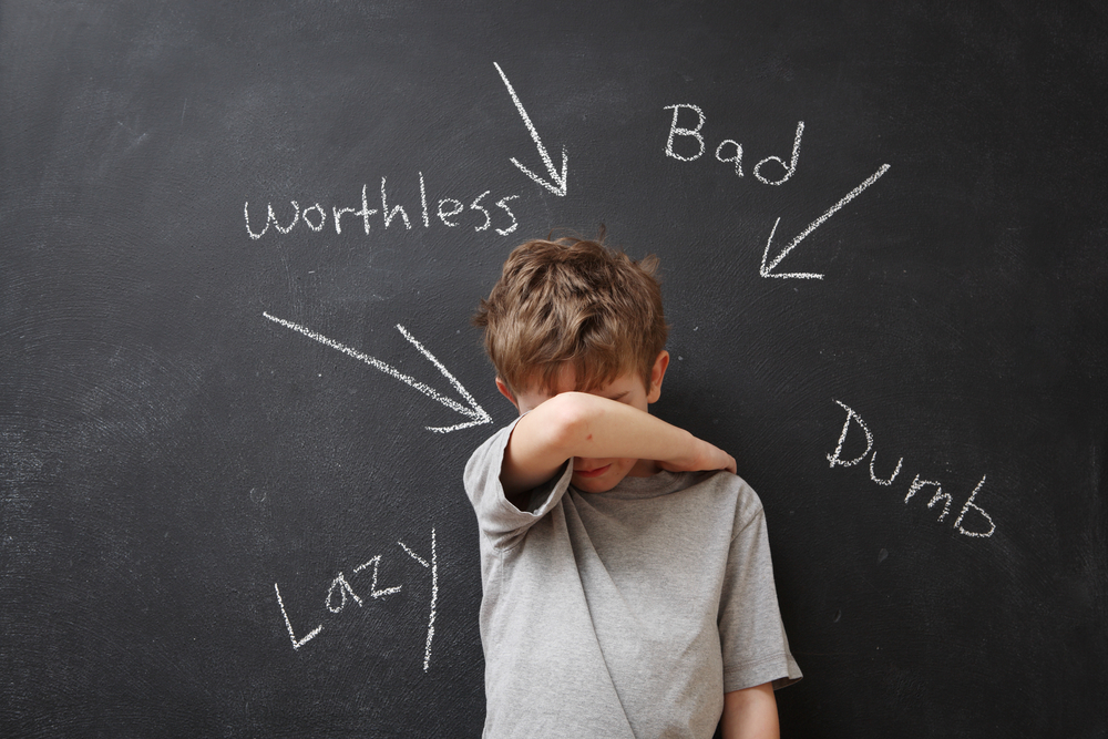 small boy with an arm covering his face and a chalkboard behind him with negative words and arrows pointed at him