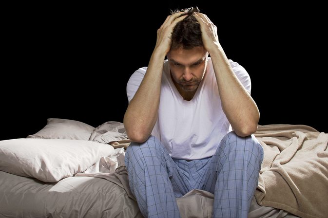 man in pajamas with his hands on his head sitting on a bed in a dark room