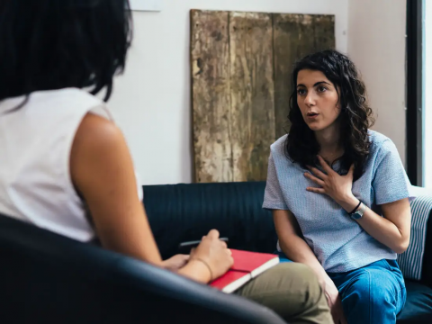 woman sitting in therapist's chair holding a red notebook and pen sitting across from a woman who is speaking with her hand over her chest and the other hand over her knees